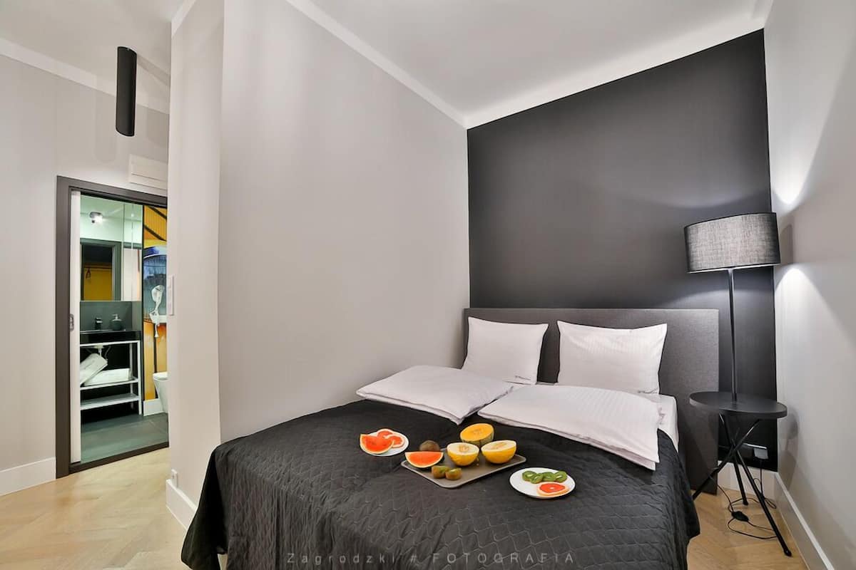 Un dormitorio moderno con un cabecero oscuro, una mesita de noche con una lámpara y una bandeja de fruta sobre la cama.