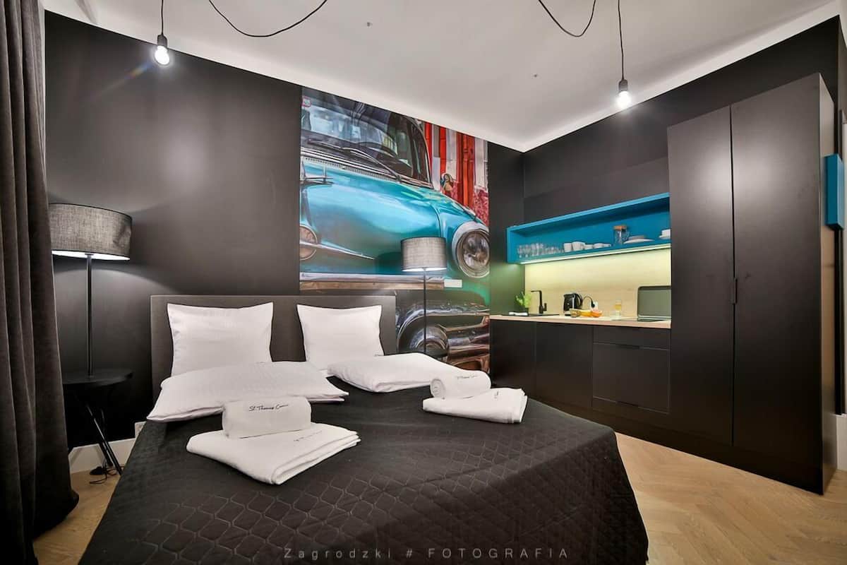 Una habitación de hotel moderna con cama, un cuadro de un coche, una kitchenette y paredes de color oscuro.