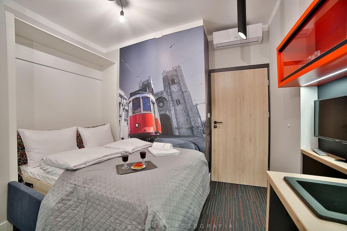 Habitación de hotel con cama, un mural en la pared de un tranvía rojo, una puerta de madera y un televisor con marco rojo.