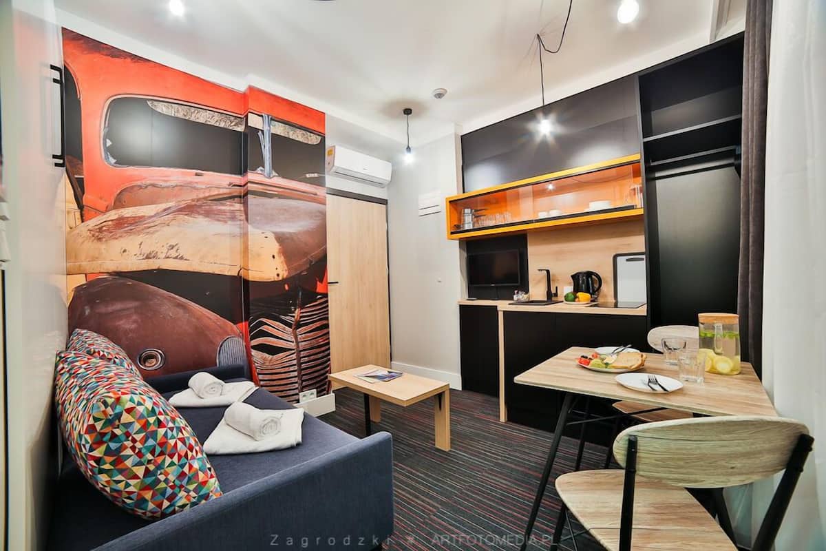 Una habitación compacta con sofá, una mesita, una mesa de comedor con sillas, una zona de cocina con refrigerador y un mural en la pared.