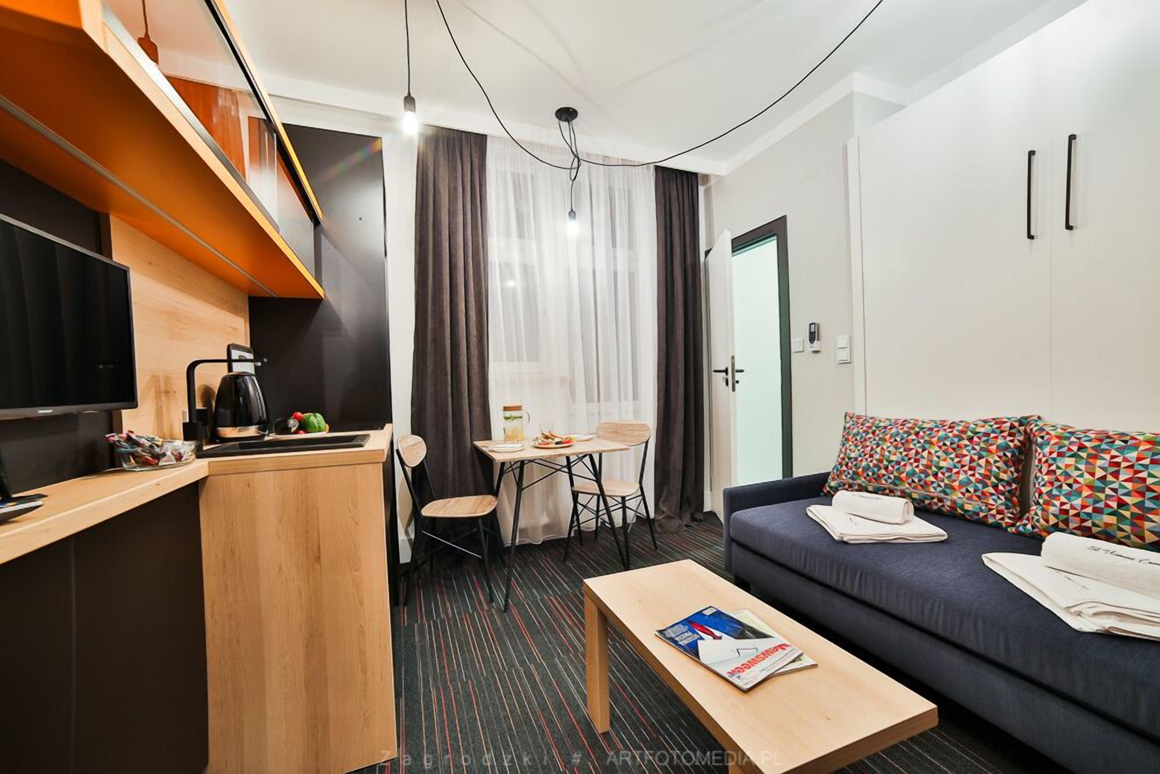 A compact hotel room with a kitchenette, a sofa, a dining table, and a TV.