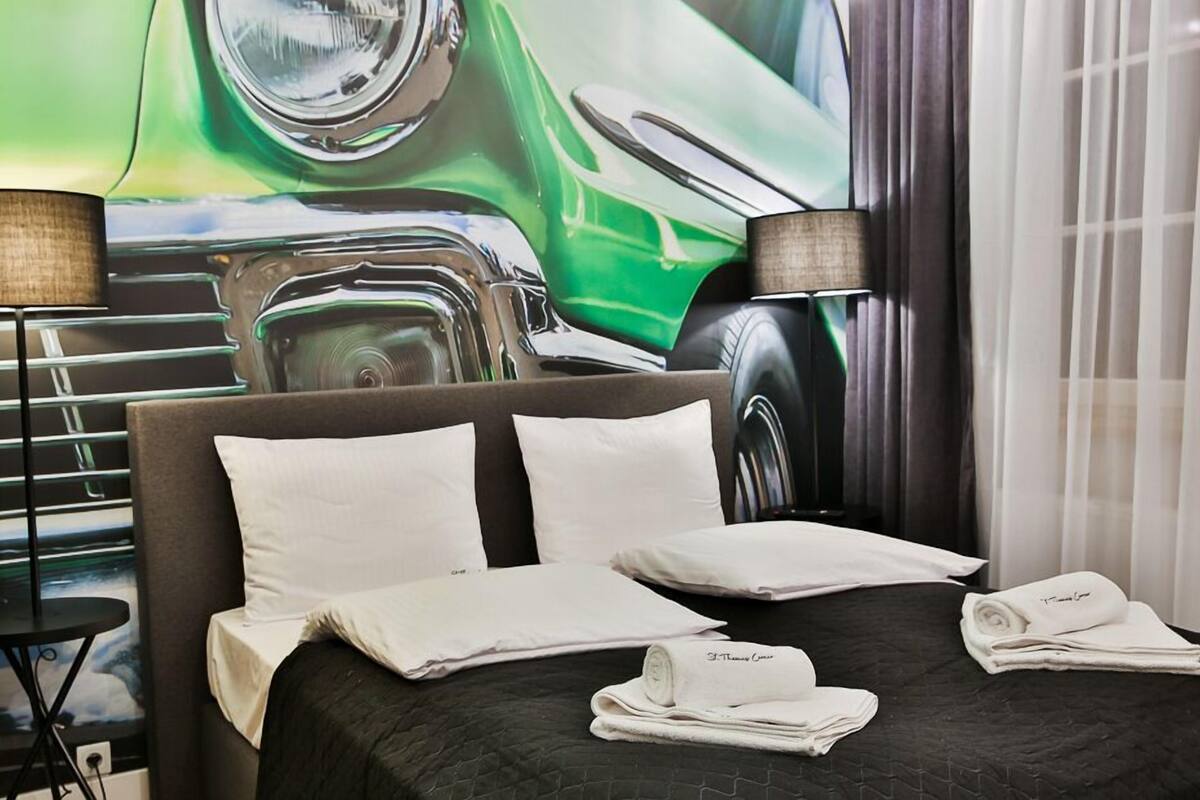 Habitación de hotel con una cama grande, dos lámparas de noche, cabecera con una imagen de coche y toallas dobladas cuidadosamente.