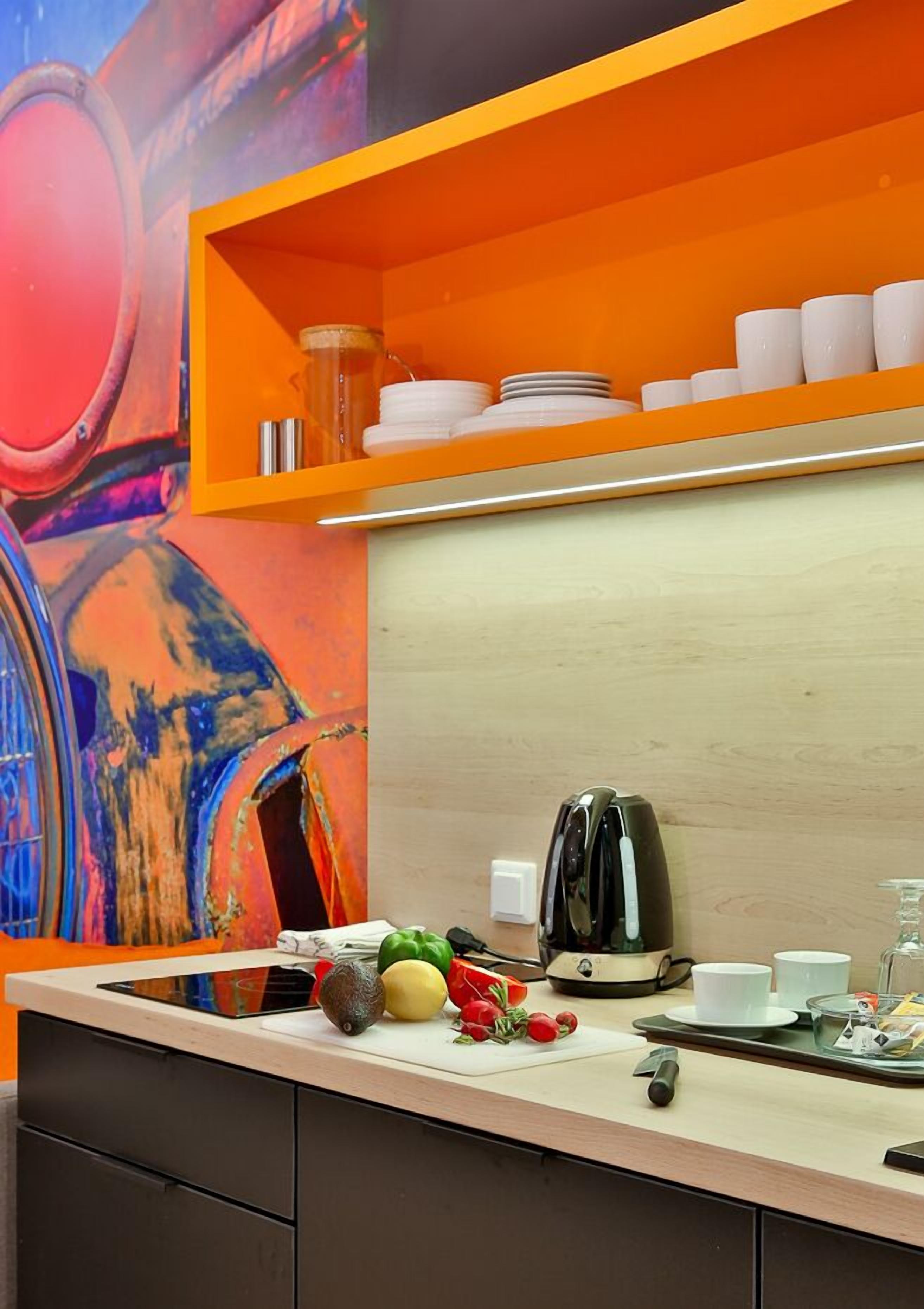 Una cocina con encimera de madera, un hervidor, una tabla de cortar con verduras y una estantería con vajilla blanca.
