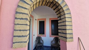 Vue intérieure d’une pièce avec un mur rose, une arche en briques, deux fenêtres et deux chaises en osier.