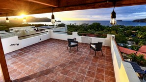Una terrazza con pavimento piastrellato, due poltrone in vimini e vista sul mare e sulle montagne al tramonto.
