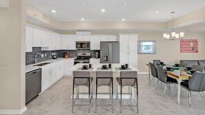 A modern kitchen with white cabinets, a central island with bar stools, stainless steel appliances, and a dining area with a table and chairs. - Modern and Spacious 2BD Condo, With Pound View, Close to Disney #3171sl306 (Kissimmee)