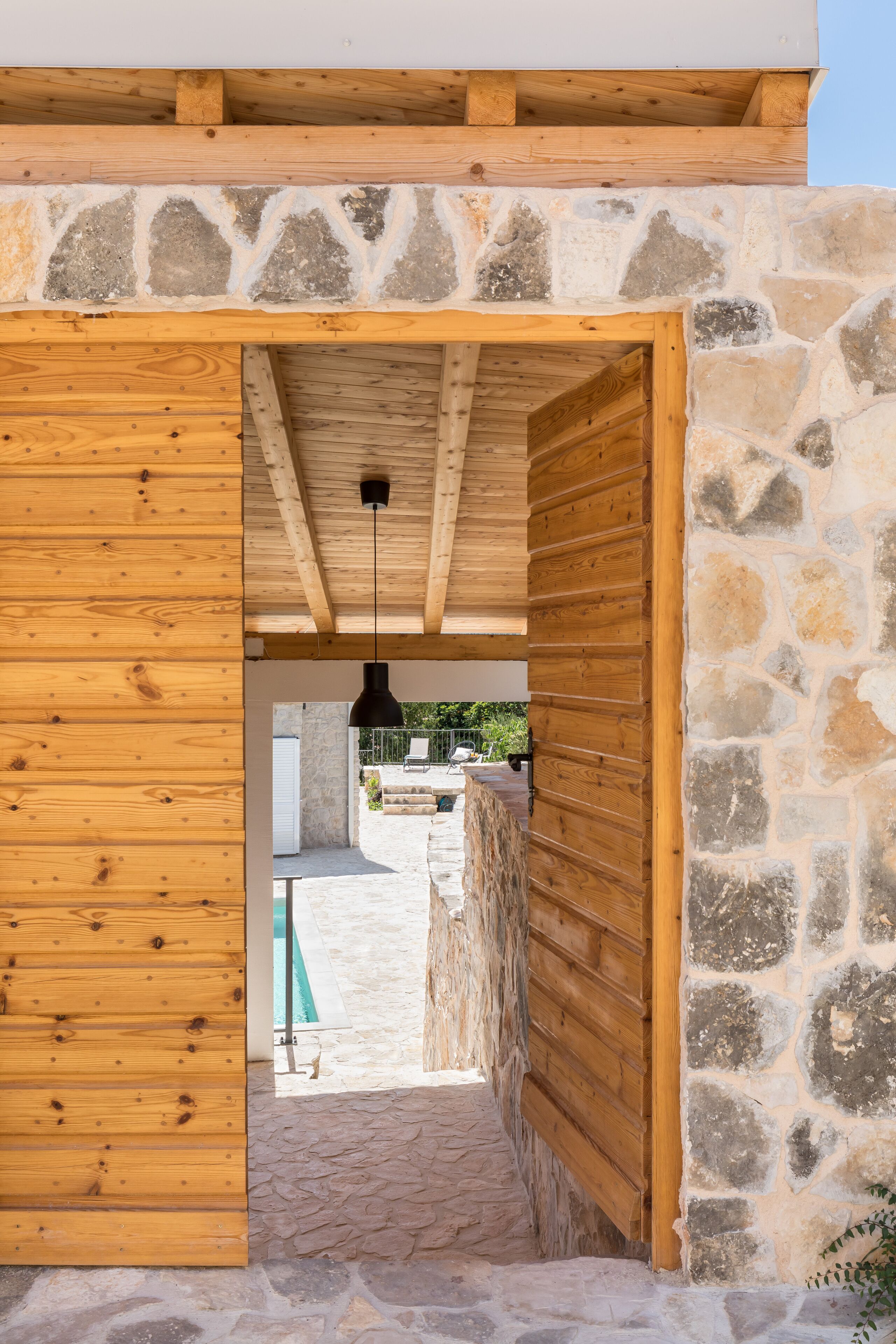A wooden door leading to a stone-walled interior with a view of an outdoor swimming pool.