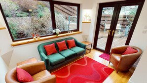 Ein gemütliches Wohnzimmer mit einem grünen Sofa, zwei braunen Ledersesseln, einem roten Teppich und großen Fenstern mit Blick auf einen Garten und eine Terrassenanlage.