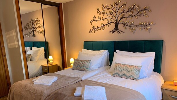 Ein Doppelbett mit smaragdgrüner Kopfteil, weißer Bettwäsche und Zierkissen. An der linken Wand befindet sich ein großer Spiegel und ein Wandbild über dem Bett.