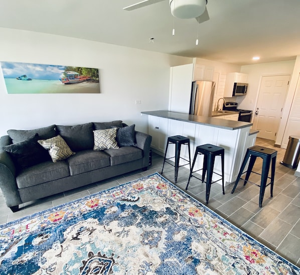 A modern living room with a gray sofa, a colorful rug, and a kitchen area with white cabinets and black stools.