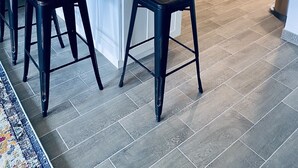 Ett modernt kök med vita skåp, ett kylskåp i rostfritt stål, en mikrovågsugn och en spis. Det finns två svarta barstolar vid köksön.