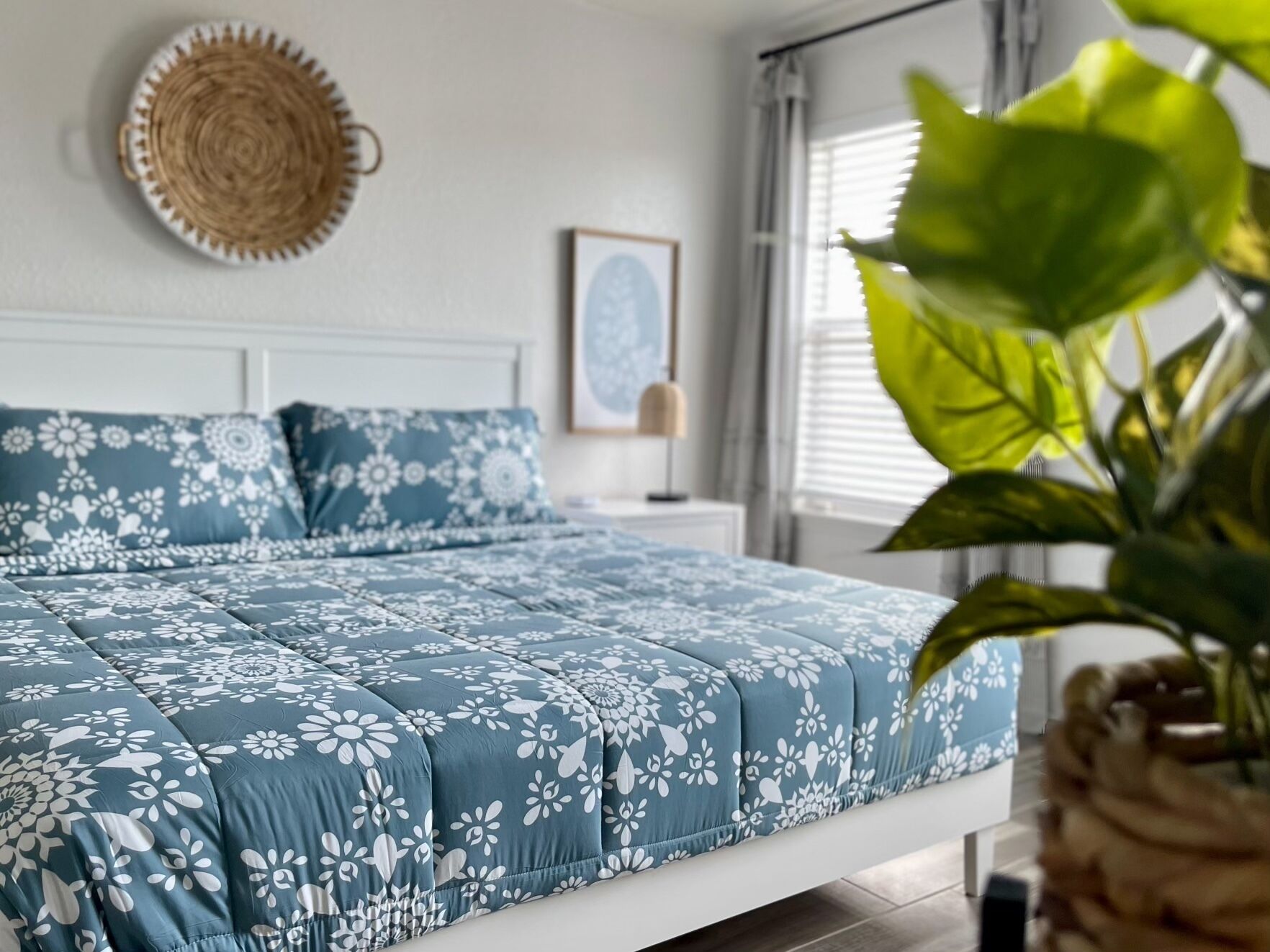 A bedroom with a bed, a bedside table, a lamp, a wall hanging, and a window with blinds.