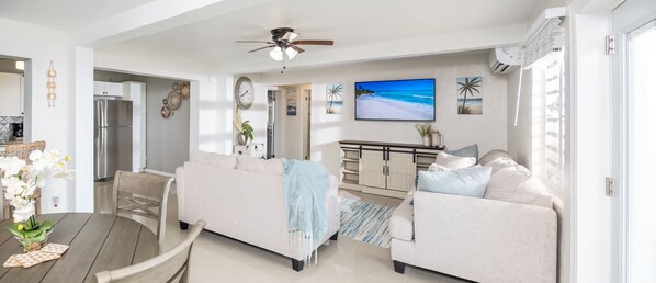 A modern living room with a sofa, a dining table with chairs, a kitchen area with a refrigerator, and a TV displaying a beach scene.