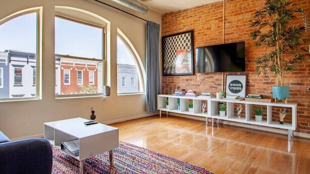A modern living room with large windows, a brick wall, a flat-screen TV, a white console table, and a colorful rug. - Artistic Abode Close to MICA & Johns Hopkins (Baltimore)