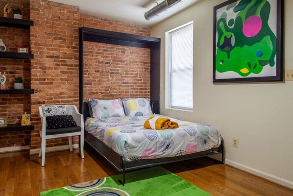 A bedroom with a brick wall, a bed with a colorful bedspread, a white chair, a green rug, and a large abstract painting on the wall.