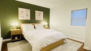 Un dormitorio con una cama, dos mesitas de noche, un aparador, un ventanal con persianas y dos obras de arte colgadas en la pared.