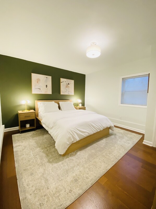 Une chambre à coucher comprenant un lit, deux tables de chevet, une commode, une fenêtre avec stores et deux œuvres d’art accrochées au mur.