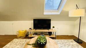Una sala de estar acogedora con un televisor sobre un soporte de madera, una planta pequeña en una mesa, una lámpara de pie y un tragaluz arriba.