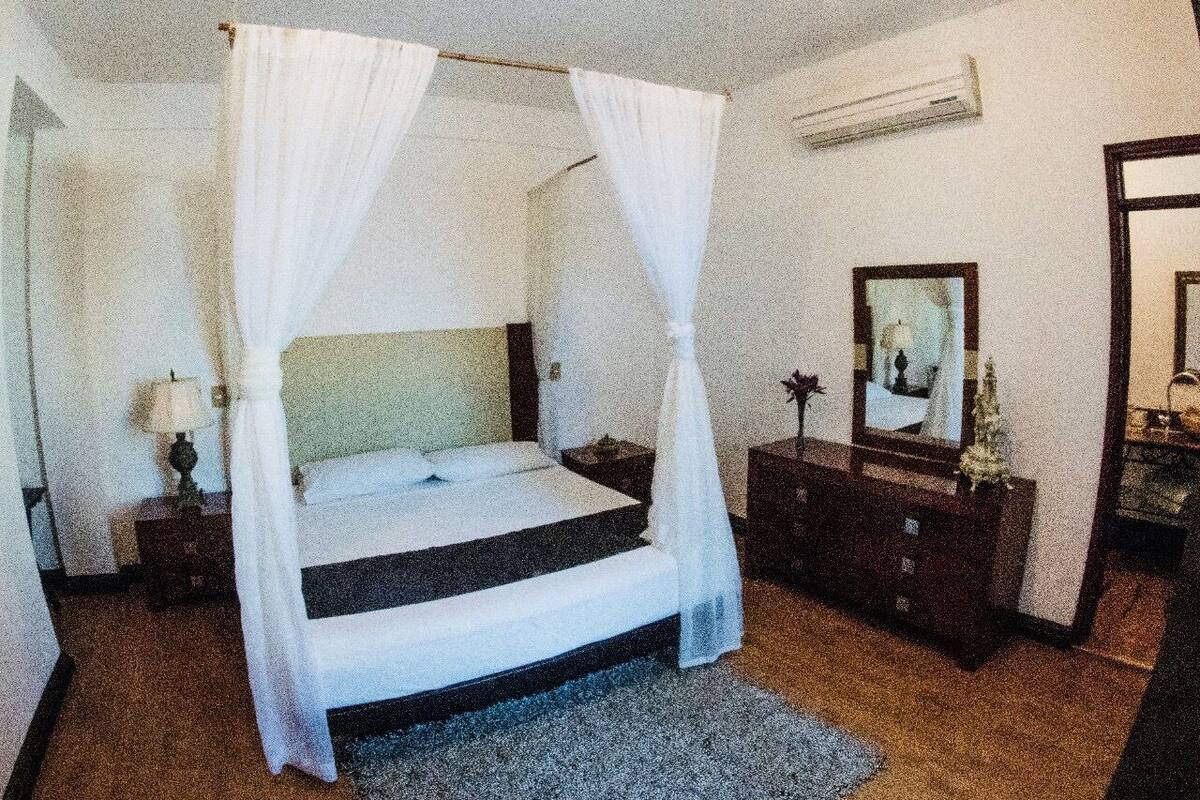 A bedroom with a four-poster bed, a nightstand with a lamp, a dresser, and a mirror.