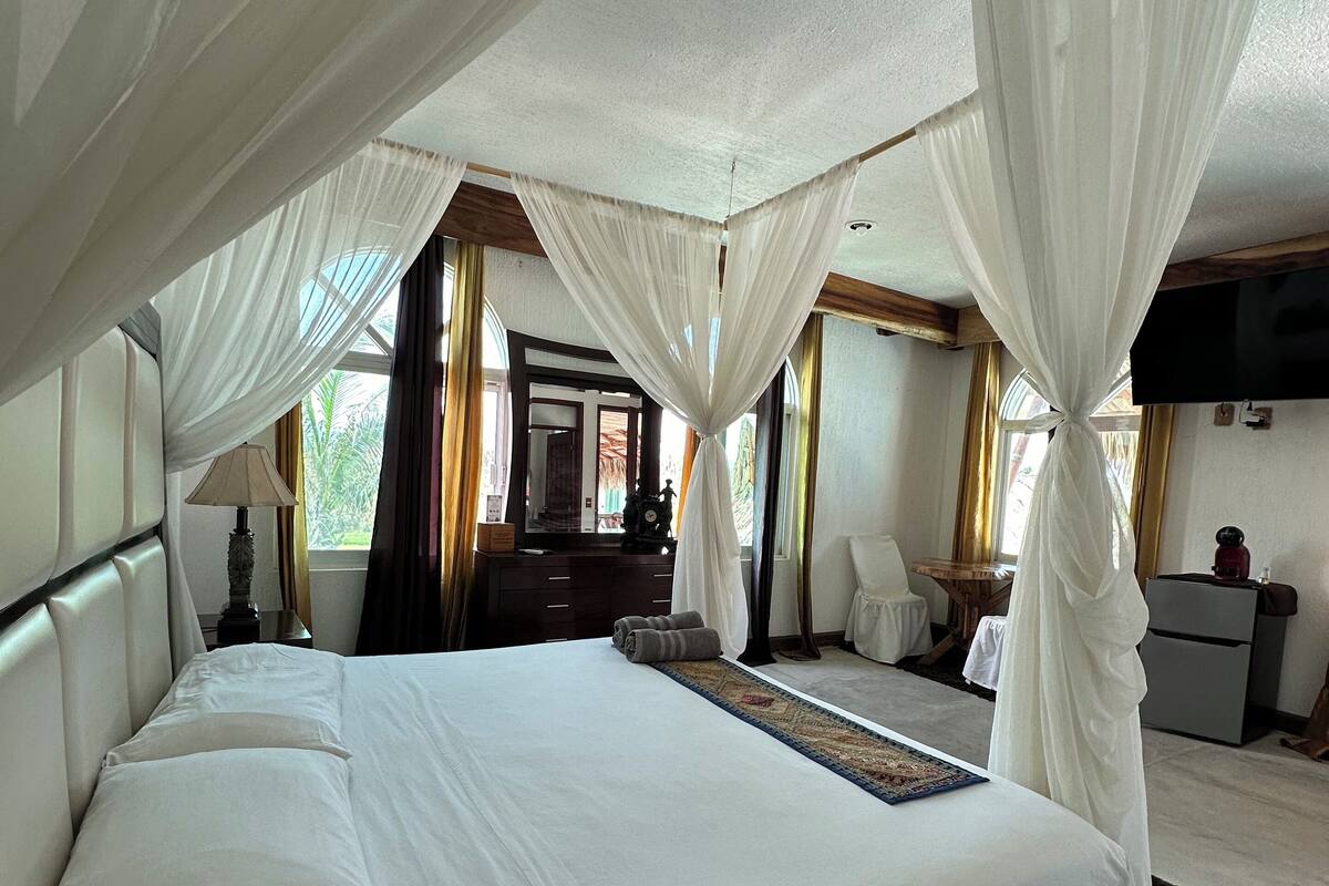A spacious bedroom with a large bed, a TV mounted on the wall, and a view of the outdoors through the windows.