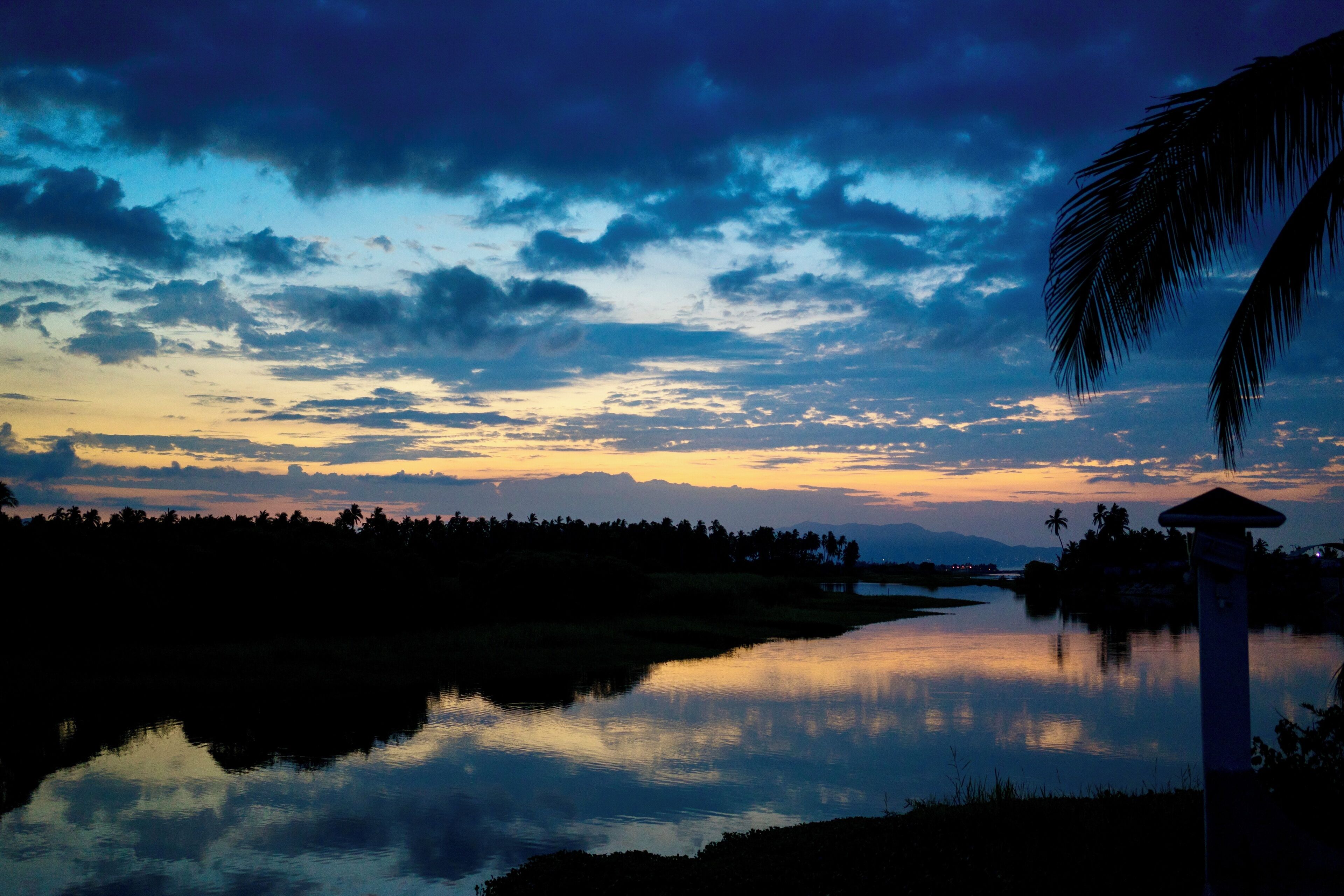 A serene sunset over a calm river with a silhouette of trees and a palm tree on the right.