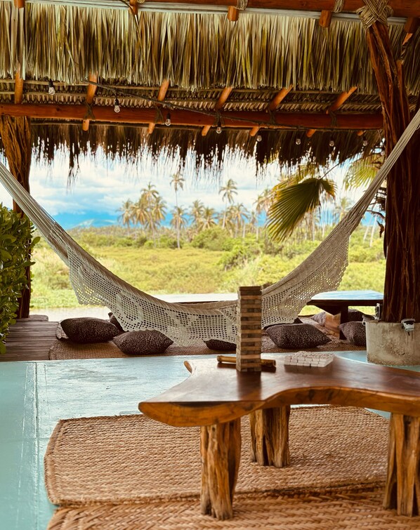 A hammock under a thatched roof, a wooden table, and a view of palm trees.