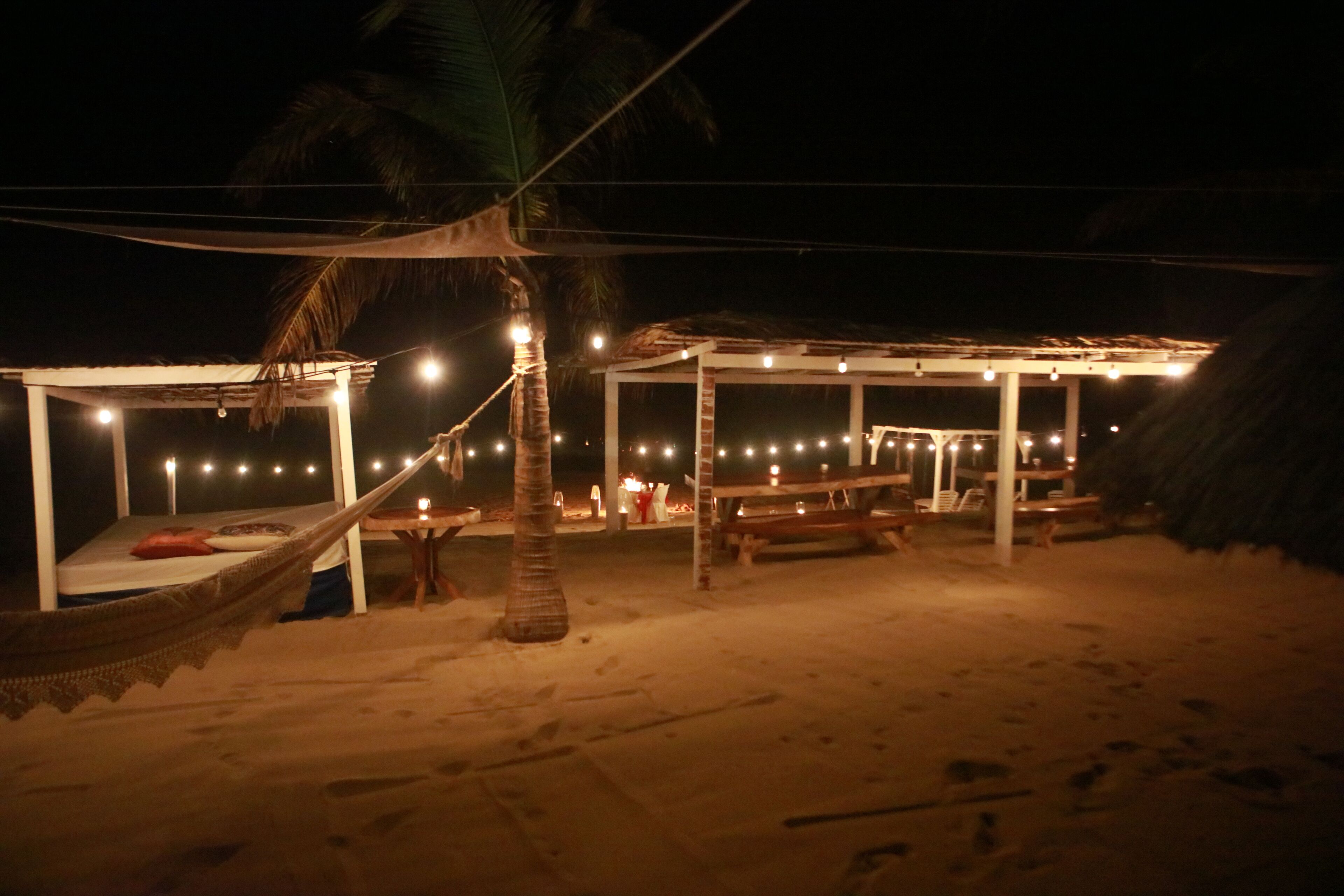Un paisaje playero con una hamaca, un espacio de descanso cubierto y luces colgantes.