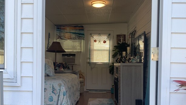 Un pasillo estrecho conduce a un dormitorio con cama, mesita de noche y lámpara. Se observa una puerta con ventanal y un pequeño porche con una silla y una planta.