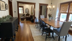 Un comedor con mesa de madera, sillas negras y una araña. Hay una alfombra en el suelo y una ventana con persianas a la derecha.
