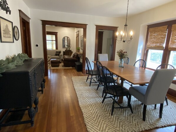 A dining area with a wooden table, black chairs, and a chandelier. There is a rug on the floor, and a window with blinds on the right.