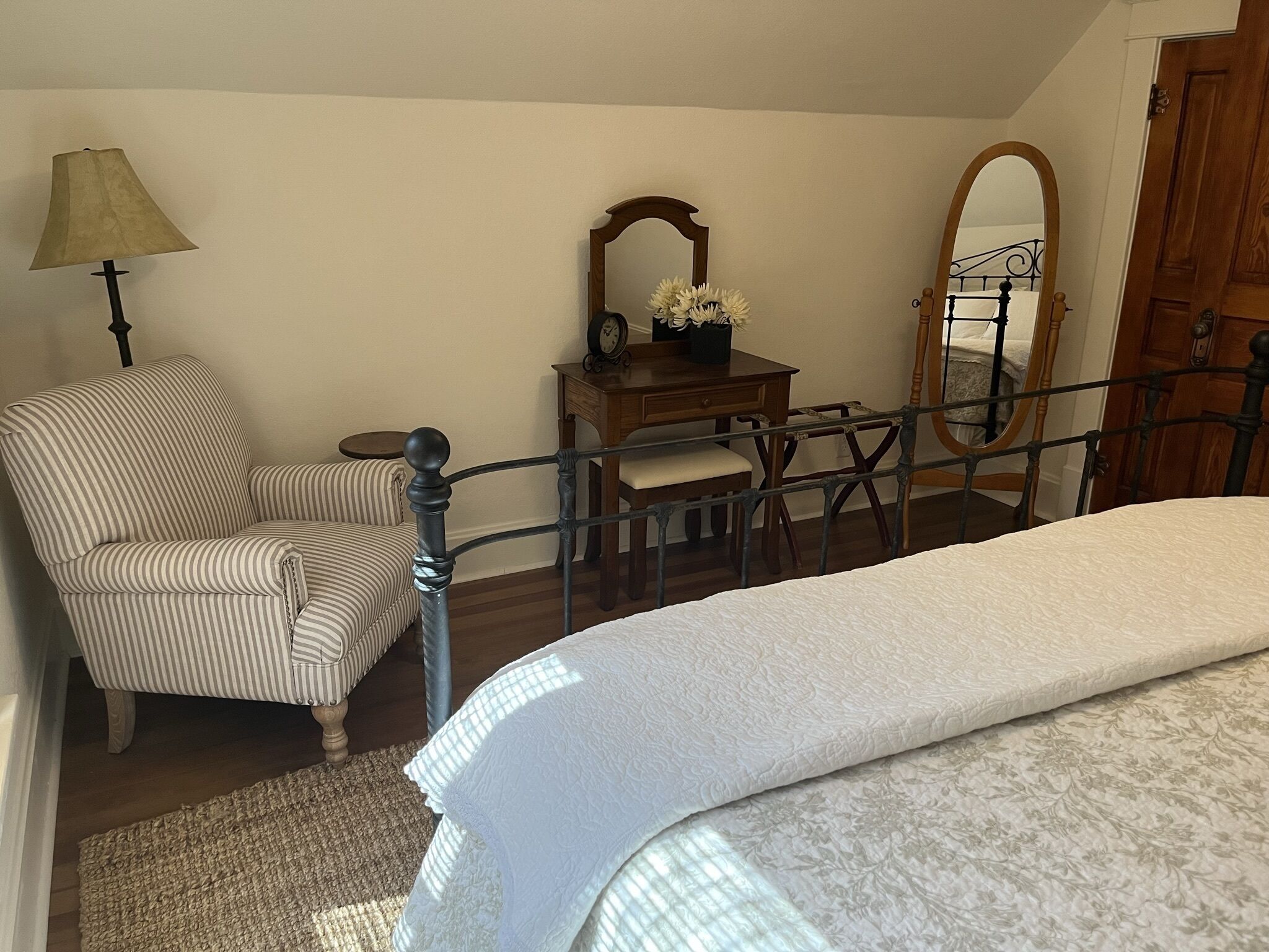 A bedroom with a bed, a chair, a desk with a mirror, and a lamp.