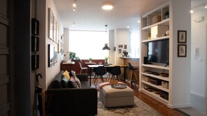 Een moderne woonkamer met een bank, een voetenbankje en een televisie op een meubel. Er is een eethoek met een tafel en stoelen, en een keuken met een televisie en planken.