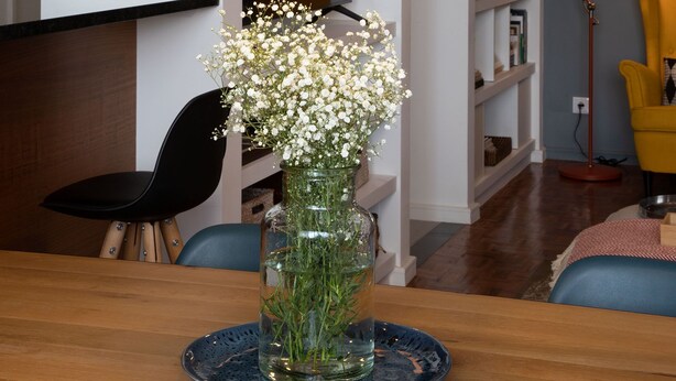 Een eetgedeelte met een tafel, stoelen, een vaas met bloemen, een hanglamp en planken met boeken en decoratieve objecten.