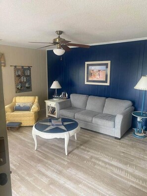 Sala de estar com sofá, poltrona, mesa de centro, ventoinha no teto e quadro na parede.