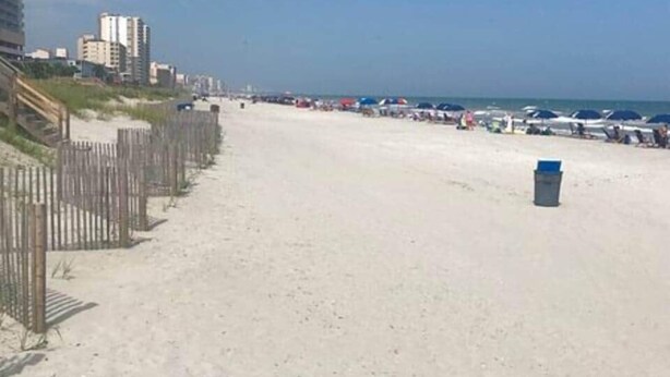 A beach with a wooden fence, a trash bin, and buildings in the background.