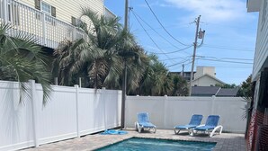 A rectangular swimming pool with a metal safety railing, surrounded by a white fence, with lounge chairs and a paved walkway.
