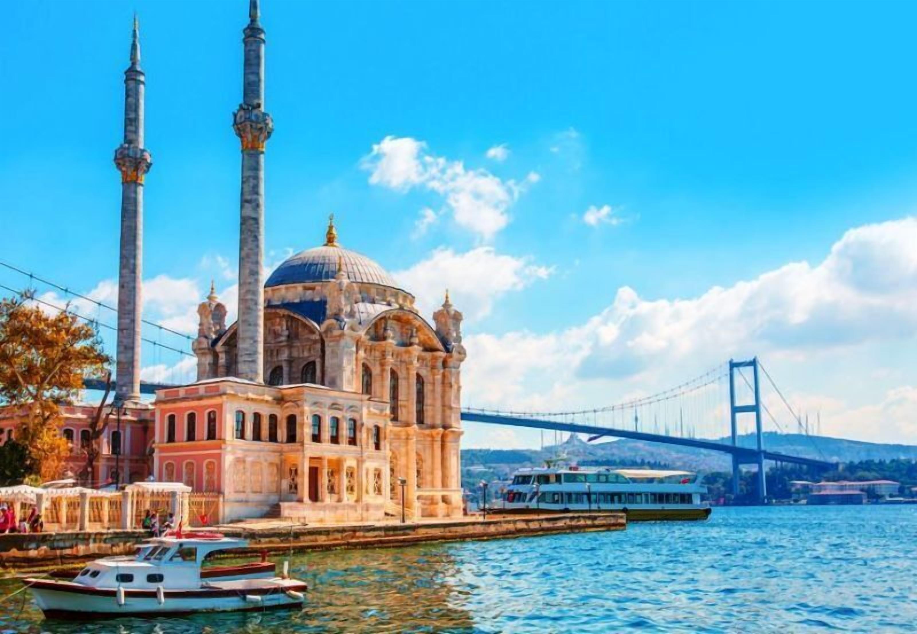A historic building with a large dome and two minarets, located near a body of water with a bridge in the background.