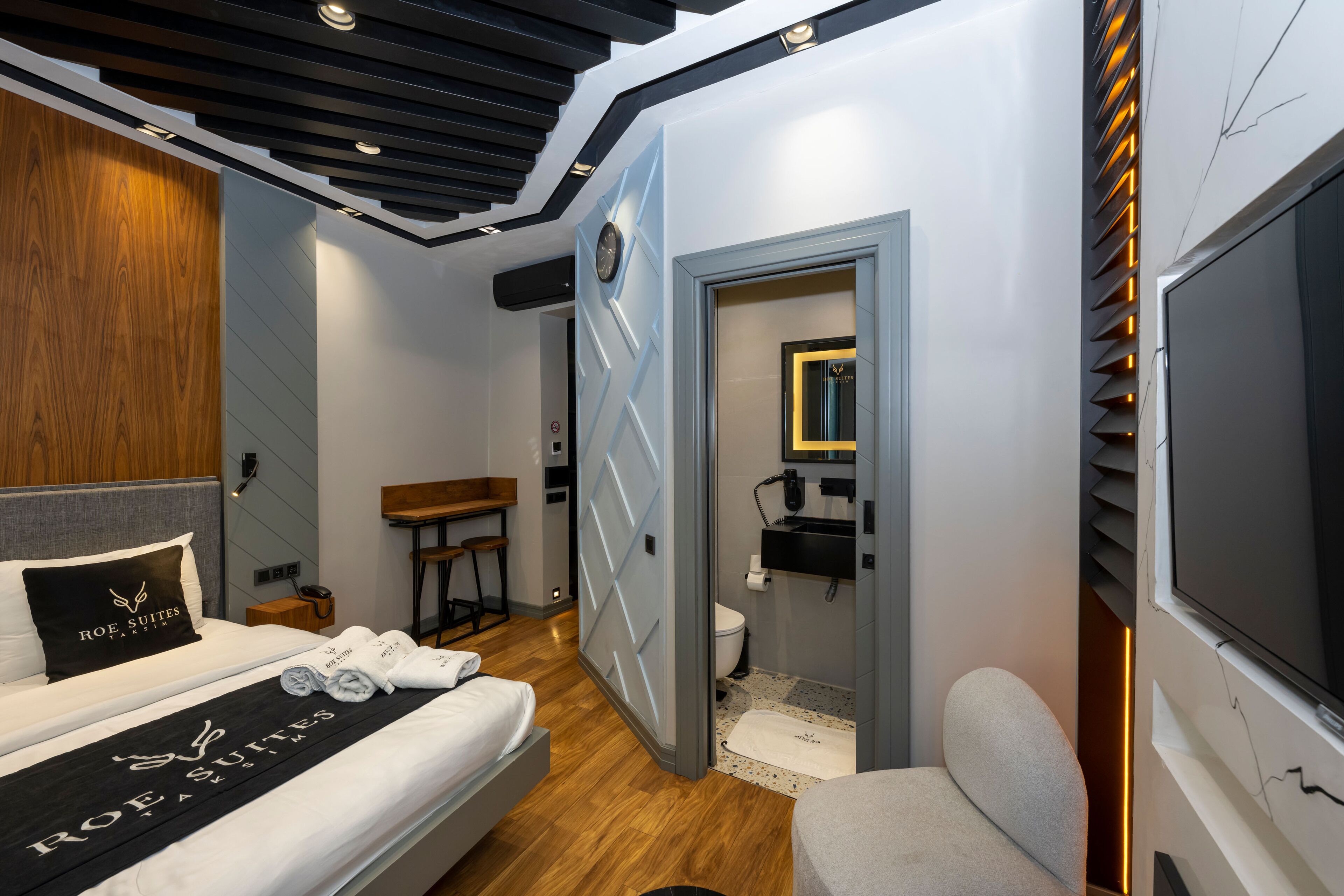 Une chambre d’hôtel moderne équipée d’un lit, d’un bureau, d’une chaise, d’une télévision et d’une salle de bain visible à travers une porte ouverte.