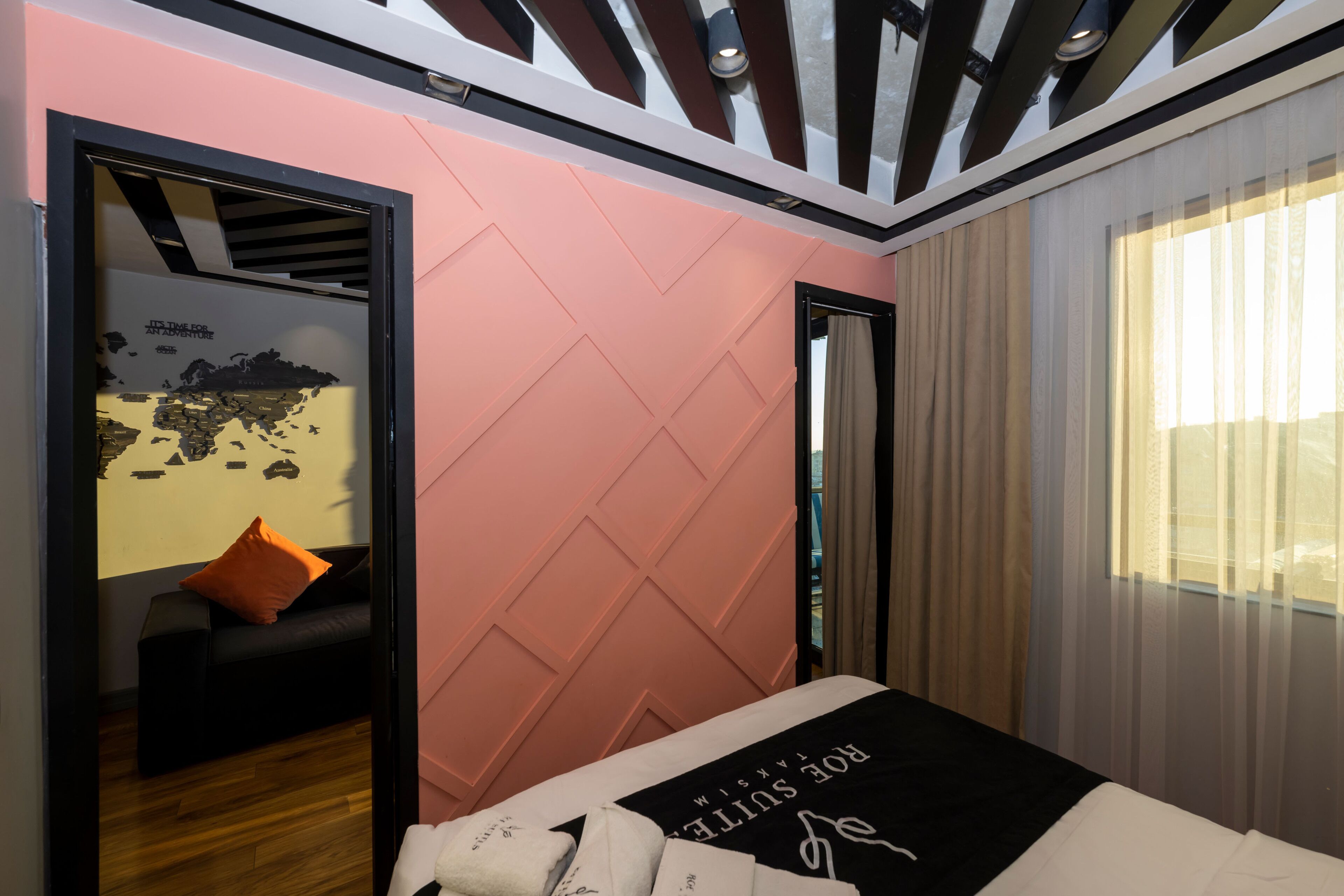 A modern hotel room with a pink accent wall, a bed with a black and white headboard, a wooden floor, a black sofa with an orange pillow, and a large window with sheer curtains.