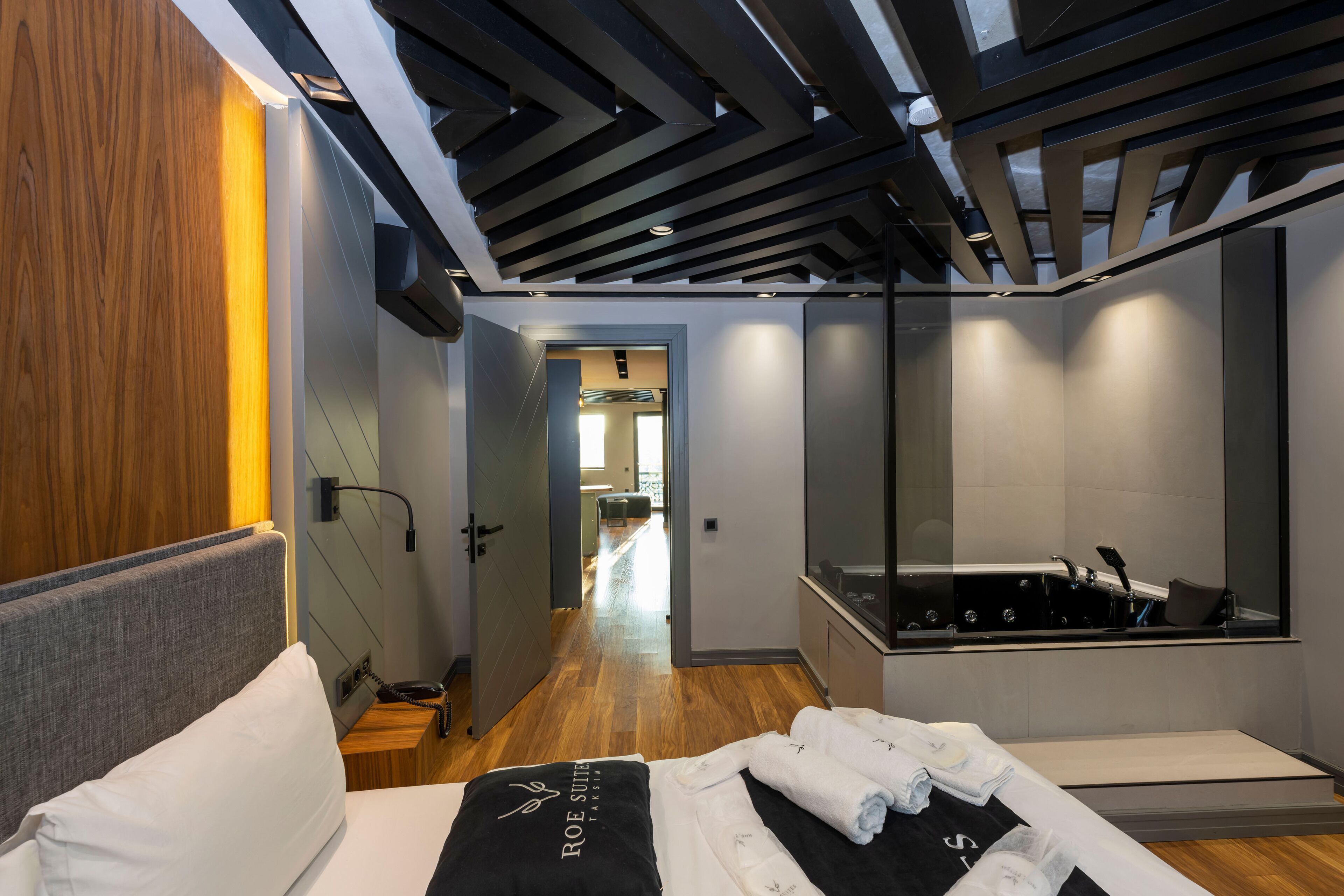 A modern hotel room with a wooden floor, a bed with a black and white towel, a large mirror, and a view into the hallway.
