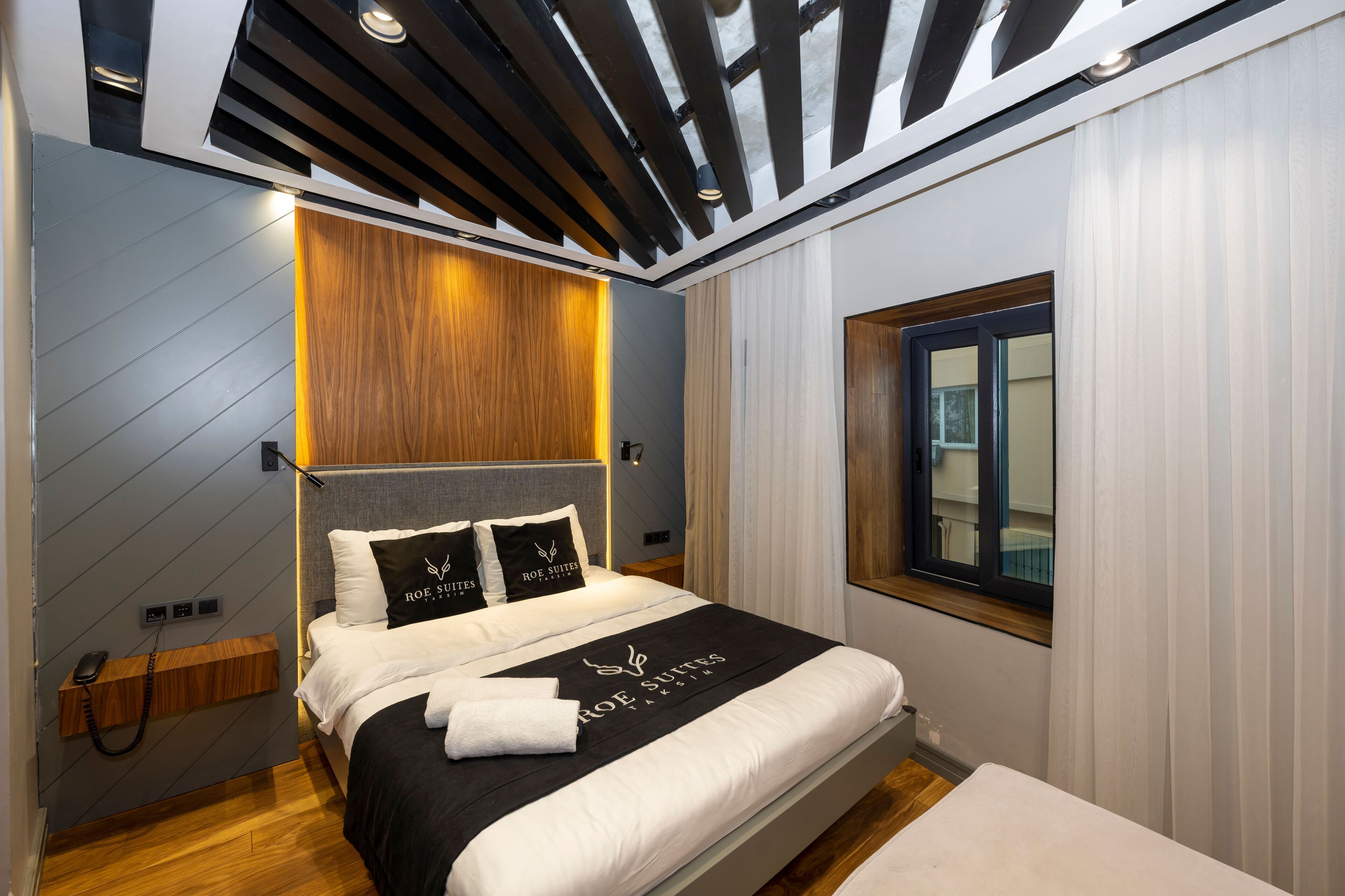 A modern hotel room with a bed, a wooden headboard, a bedside telephone, and a window with curtains.