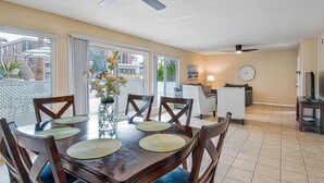 Un coin repas avec une table en bois, des chaises et un vase de fleurs. On y trouve également un ventilateur de plafond, une horloge et un téléviseur posé sur un meuble.