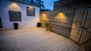 Una terraza de madera al aire libre con una pared blanca y paneles metálicos decorativos, una valla de madera y una planta en maceta.