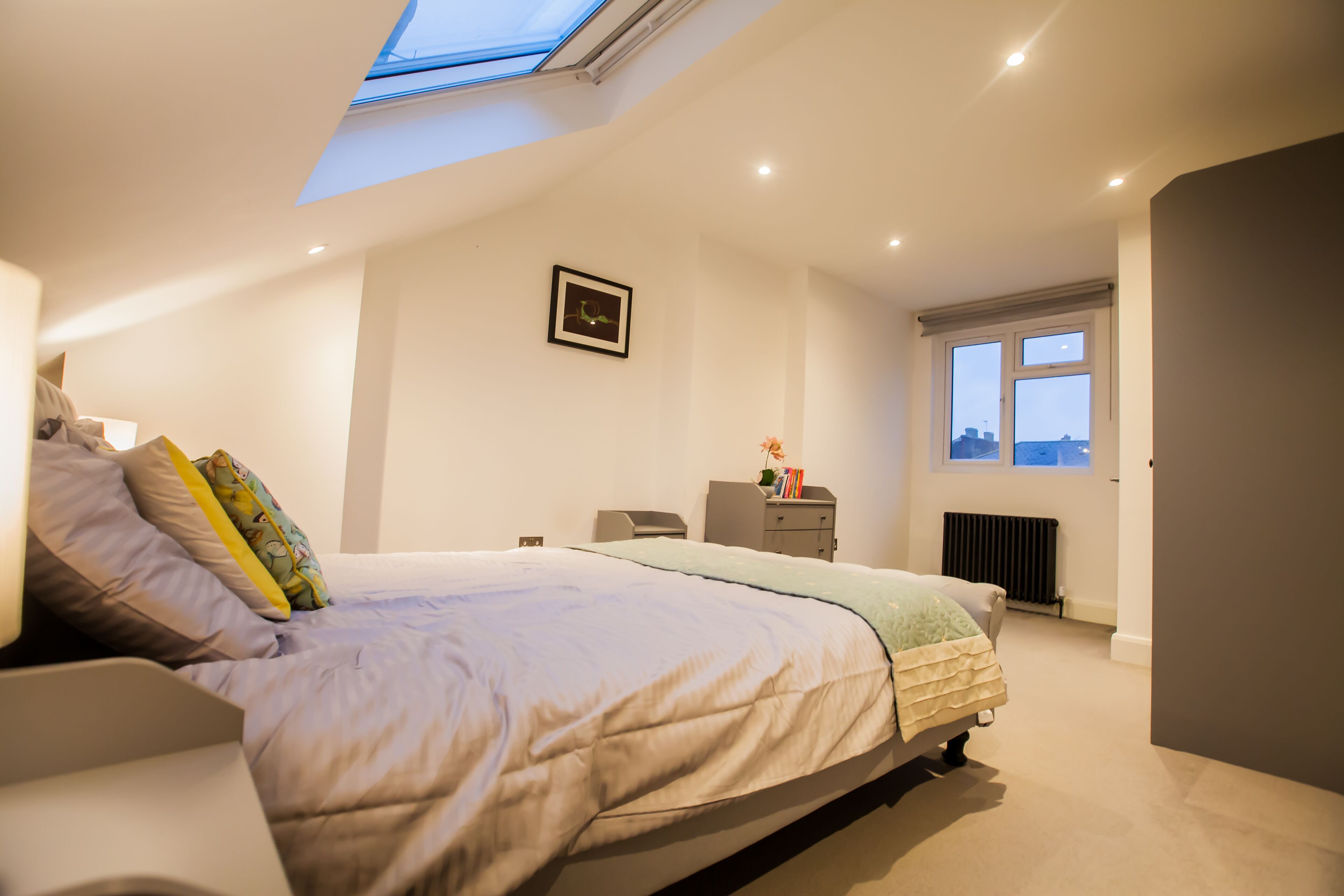 A bedroom with a skylight, a bed with multiple pillows, a bedside table, a radiator, and a window with a view.