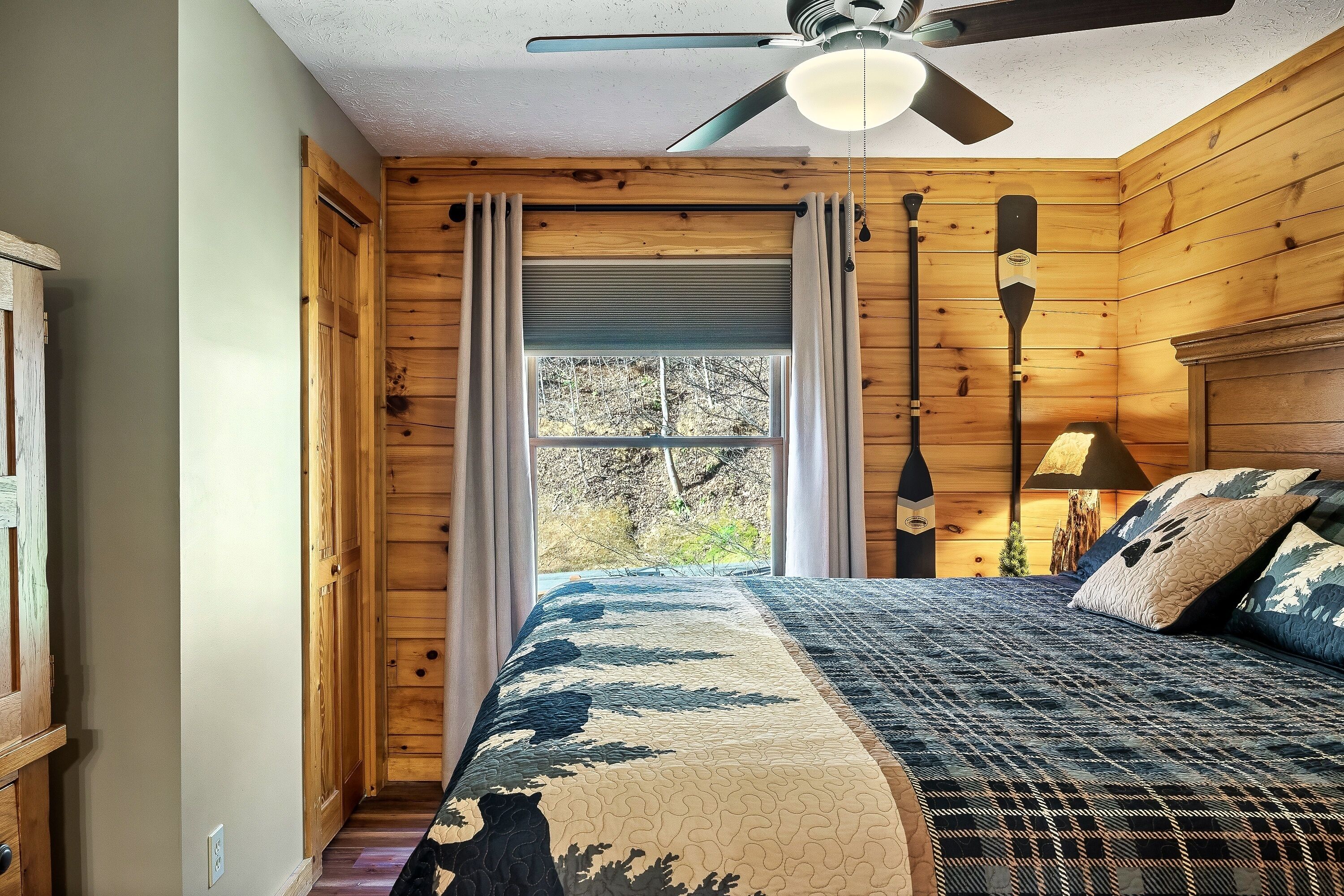 A wooden cabin bedroom with a bed, a ceiling fan, a window with curtains, and a lamp.