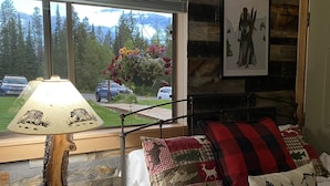 Een slaapkamer met een bed, een lamp en een raam met uitzicht op een bos en bergen.