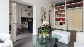 Ein modernes Wohnzimmer mit einem Glastisch, weißen Sofas, einem Bücherregal und einer Küchenzeile im Hintergrund.