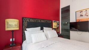 Ein Schlafzimmer mit roter Wand, einem Bett mit weißer Bettwäsche, einem Nachttisch mit Lampe und einem an der Wand montierten Fernseher.