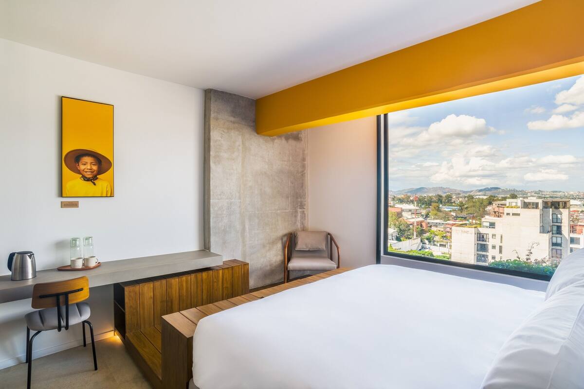 A modern hotel room with a large bed, a desk with a chair, a coffee station, and a city view through a large window.