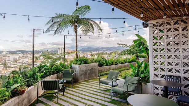 A rooftop terrace with string lights, green chairs, a table, and a view of a cityscape and mountains.